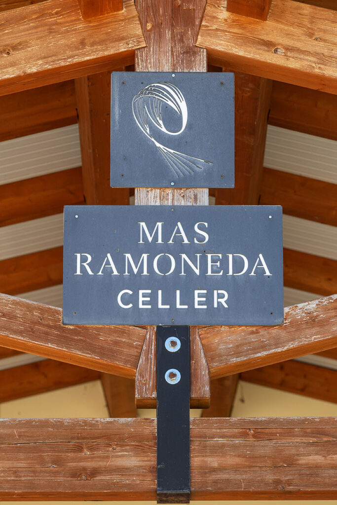Logotip al cartell del celler Mas Ramoneda, sota un porxo de fusta amb bigues de fusta, per visitar el celler.